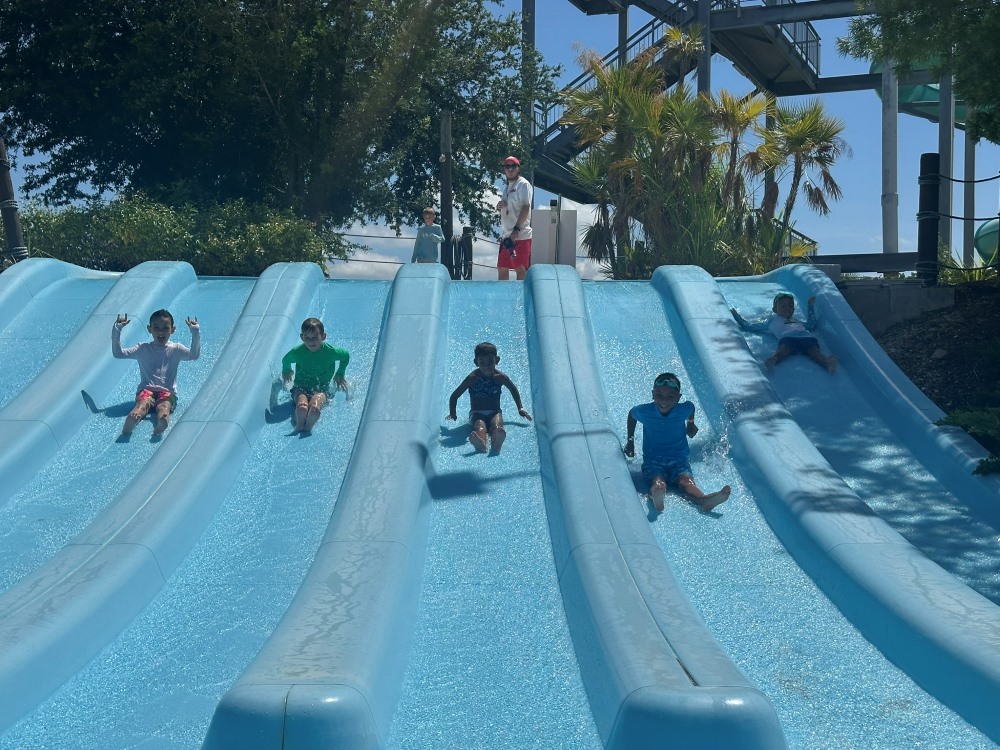 Five children race down parallel blue water slides while a lifeguard watches from the top platform.